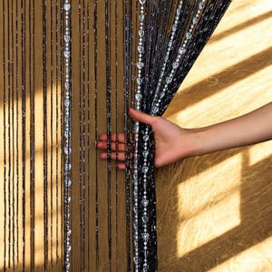 Puede incluir: Cortina de cuentas negras con detalles plateados en forma de corazón y redondos. Una mano sostiene la cortina abierta, revelando una superficie de madera. Los hilos verticales de cuentas crean un efecto brillante. Accesorio decorativo.
