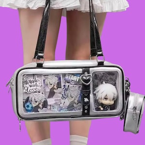 Puede incluir: Bolso rectangular plateado con asas negras y una pequeña bolsa a juego. La bolsa presenta compartimentos transparentes que muestran ilustraciones de estilo anime y una pequeña figura. El texto "made with love" y "START" son visibles.