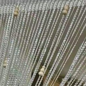 May include: A decorative curtain with strands of silver-colored, chain-like material. The strands are adorned with small, off-white, gourd-shaped beads. The curtain hangs in vertical lines, creating a shimmering effect.