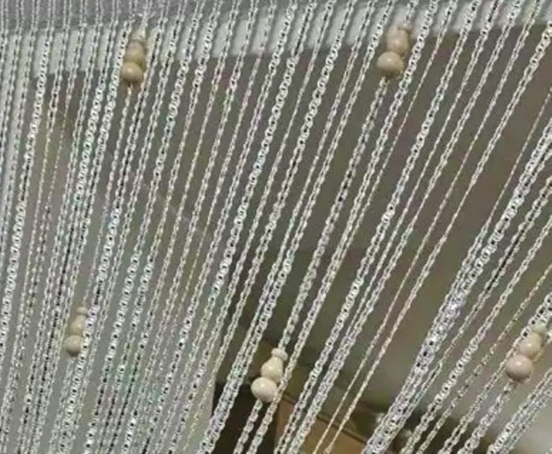 May include: A decorative curtain with strands of silver-colored, chain-like material. The strands are adorned with small, off-white, gourd-shaped beads. The curtain hangs in vertical lines, creating a shimmering effect.