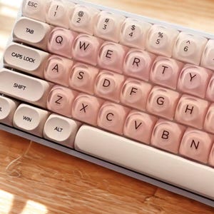 Könnte beinhalten: Nahaufnahme einer weißen und rosa mechanischen Tastatur. Die Tastenkappen sind ein Farbverlauf aus Rosa und Weiß, wobei die Buchstaben und Symbole in einer dunkleren Farbe gedruckt sind. Die Tastatur liegt auf einer Holzoberfläche.