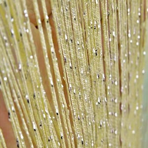 Pode incluir: Close-up de uma cortina amarela com detalhes metálicos prateados. A cortina é feita de inúmeros fios verticais, criando um efeito brilhante. Os fios estão próximos e os detalhes prateados são distribuídos irregularmente.