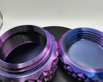 May include: A purple and blue iridescent metal container with a textured, geometric design. The container is open and the lid is sitting next to it.