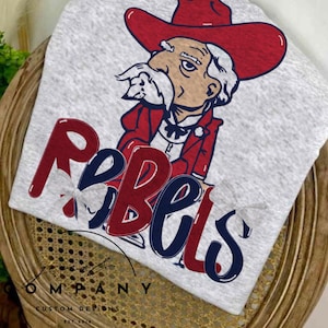 May include: A light gray t-shirt featuring a cartoon illustration of a man in a red cowboy hat and jacket. The word "Rebels" is printed in red and blue. The shirt is displayed on a wooden chair.