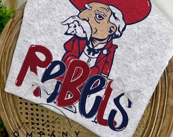 May include: A light gray t-shirt featuring a cartoon illustration of a man in a red cowboy hat and jacket. The word "Rebels" is printed in red and blue. The shirt is displayed on a wooden chair.