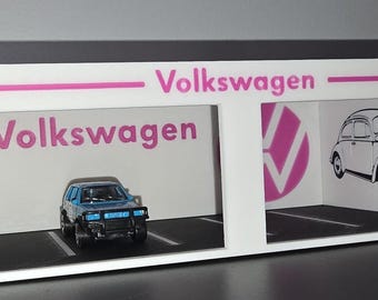 Puede incluir: Una vitrina blanca con la palabra "Volkswagen" en rosa. Un SUV de juguete azul y negro está estacionado en una plaza de aparcamiento. Una ilustración en blanco y negro de un clásico Volkswagen Beetle está a la derecha.