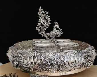 Puede incluir: Plato de servir plateado ornamentado con una figura de pavo real en el centro. El plato presenta detalladas tallas florales y de figuras alrededor del borde e incluye cuatro cuencos pequeños. El plato está sostenido por tres pies decorativos.