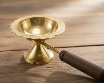 Puede incluir: Una lámpara de aceite de latón dorado con borde festoneado y mango de madera. La lámpara está sobre una superficie de madera. La lámpara probablemente se utiliza con fines religiosos o decorativos.