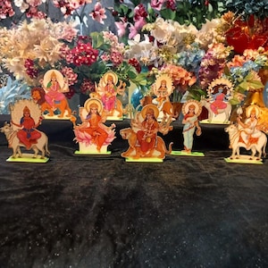 May include: A selection of colourful wooden figurines illustrating various deities. Each figure is mounted on a green base and features intricate details, vibrant colours, and traditional attire. The background includes a display of fresh flowers.