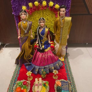 May include: A traditional Indian wedding ceremony scene with three dolls dressed in colorful clothing. The dolls are sitting on a red velvet platform with gold trim. There are several small trays with fruit and flowers on the platform.