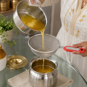 May include: A stainless steel mesh strainer with a red handle is positioned over a stainless steel container. A golden liquid is being poured from a stainless steel saucepan through the mesh strainer into the container below.