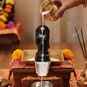 May include: A religious ceremony with a black statue being bathed in white liquid from a small brass pot. The statue is on a small wooden table with a red patterned cloth, surrounded by marigold flowers and incense sticks.