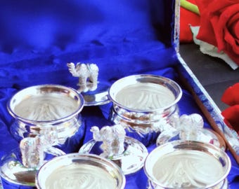 Puede incluir: Un juego de cuencos plateados con tapas, cada uno coronado con una pequeña figura de elefante. Los cuencos están dentro de una caja forrada de azul con un exterior estampado. El juego se exhibe sobre una superficie oscura.