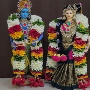 May include: A pair of handmade dolls dressed in traditional Indian clothing. The male doll is blue with a peacock feather headdress and holds a flute. The female doll is wearing a gold and green sari with a red and pink blouse. Both dolls are adorned with flower garlands.