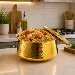Könnte beinhalten: Eine goldene Servierschüssel aus gehämmertem Metall, gefüllt mit Biryani, einem Reisgericht mit Hühnchen und Kräutern. Der Deckel ist teilweise geöffnet und gibt den Inhalt frei. Die Schüssel steht auf einer Marmorarbeitsplatte.