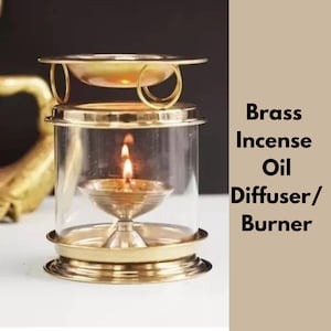 Può includere: Un diffusore/bruciatore di olio per incenso in ottone con una candela accesa all'interno di un involucro di vetro. Il bruciatore ha una base rotonda e un vassoio superiore, con il testo "Brass Incense Oil Diffuser/Burner" sul lato.