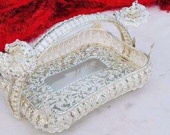 May include: Ornate silver-colored serving tray with a rectangular shape and intricate floral and scrollwork patterns. The tray has a handle and decorative floral accents on each end. The background includes a red, textured fabric.