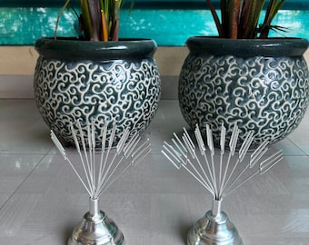 May include: Two silver-colored card holders with a conical base and radiating wire arms. The holders are in front of two dark green ceramic pots with a white swirling pattern, each containing a green plant.