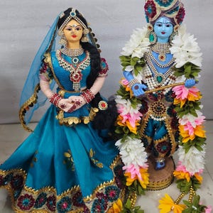 May include: Two decorative dolls representing Radha and Krishna. The female figure wears a blue dress with gold trim and jewelry. The male figure has blue skin, wears a turban, and is adorned with a large garland of white, pink, and yellow flowers.