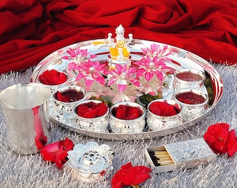 May include: A silver tray with a silver diya, eight silver bowls, and pink and white flowers. The tray is on a white fluffy surface.