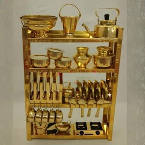 Puede incluir: Un juego de cocina en miniatura dorado sobre una estantería de varios niveles. El juego incluye un cuenco, un cubo, una tetera, ollas, platos, cucharas, tenedores, cuchillos, tazas y una estufa. Las palabras "HAPPY HOME" son visibles.