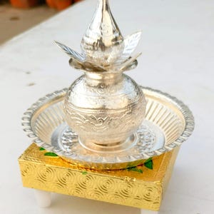 Op de afbeelding: Een zilveren kalash, een traditionele Indiase ceremoniële pot, met een deksel en een decoratieve basis. De kalash staat op een gouden standaard met groene accenten.