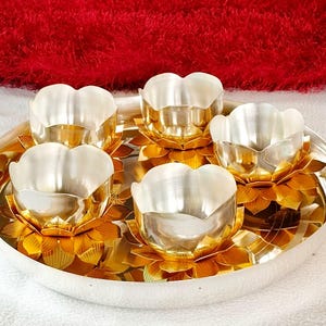 May include: A circular silver-toned tray holding five silver-toned bowls, each resting inside a gold-toned metal lotus flower base. The bowls have a smooth, reflective interior surface, while the lotus petals feature detailed, etched line textures.