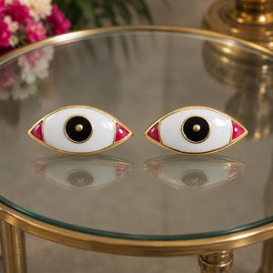 May include: A pair of eye-shaped decorative objects featuring a white sclera, a black iris with a central gold dot, and small red triangular accents at the corners. Each piece is outlined in a gold-toned metal frame with a smooth, glossy finish.