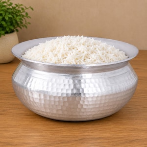 Pode incluir: Uma tigela de metal prateado cheia de arroz branco. A tigela tem uma textura martelada no exterior e uma borda larga. O arroz é fofo e enche a tigela até o topo. A tigela está sobre uma superfície de madeira.