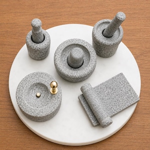 Puede incluir: Un conjunto de utensilios de cocina de granito gris, que incluye un mortero y una maja, una pequeña piedra de moler y una piedra redonda con un pomo dorado. Los artículos están dispuestos sobre una bandeja circular blanca.
