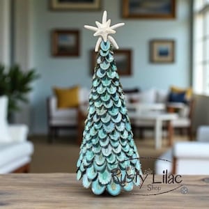 May include: A decorative Christmas tree with a coastal theme. The tree is cone-shaped, covered in teal and white shell-like elements, and topped with a white starfish. The base is a light teal color. The Rusty Lilac Shop logo is visible.