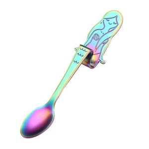 May include: A rainbow-colored metal spoon featuring a mermaid-shaped handle. The handle is bent to allow the spoon to hook onto the edge of a cup. The mermaid design includes engraved wave patterns and a stylized face with closed eyes.