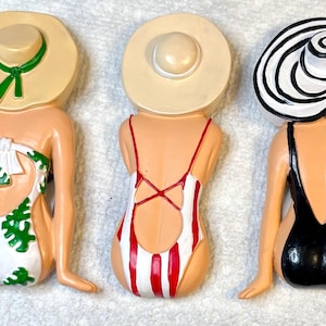 May include: Three decorative figurines of women in swimsuits and sun hats. The swimsuits are white with green leaf patterns, red and white stripes, and solid black. The hats are beige, and black and white.