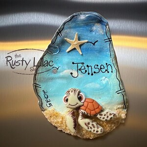 May include: A decorative, hand-painted seashell magnet featuring a cartoon sea turtle on a sandy beach with a blue sky background. The magnet includes the name "Jensen" and a starfish. The Rusty Lilac Shop logo is visible.
