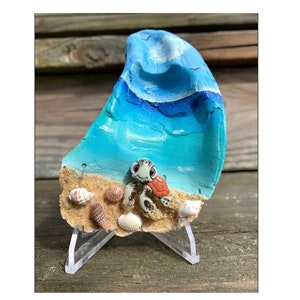 May include: A decorative oyster shell art piece featuring a painted ocean scene with a blue wave and turquoise water. The shell includes a sandy beach with seashells and a turtle family. The art piece is displayed on a clear acrylic stand.