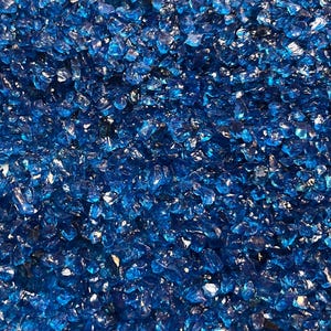 May include: Close-up of a pile of crushed, reflective, blue glass. The glass pieces are irregularly shaped and vary in size, creating a textured surface. The color is a deep, vibrant blue with highlights.