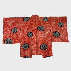 Puede incluir: Un kimono rojo con un patrón de teñido anudado blanco y rojo. El kimono presenta diseños geométricos negros y grises. La prenda está extendida, mostrando su forma y diseño completos.