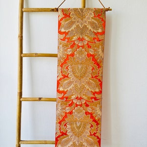 May include: A long, orange fabric wall hanging with a gold floral pattern. The fabric is hanging on a bamboo ladder.