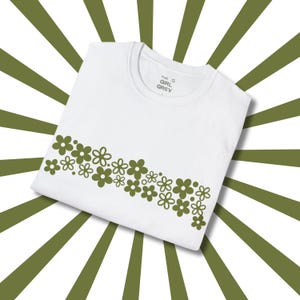 May include: White t-shirt with a row of green flowers printed across the chest. The text "THE GIRL GREY" is printed on the shirt.