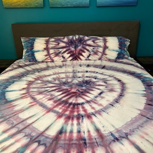 May include: A tie-dye bed set featuring a spiral design in shades of white, purple, and blue. The set includes a fitted sheet, a flat sheet, and two pillowcases. The bedding is on a bed with a gray headboard.