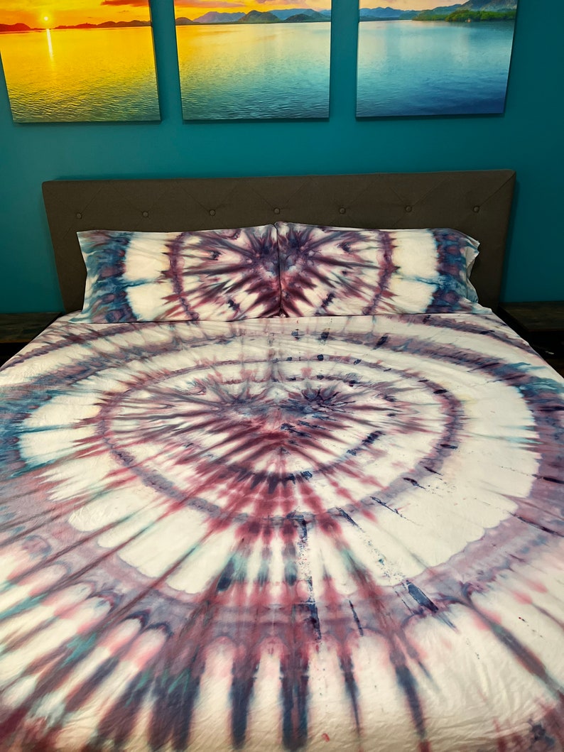 May include: A tie-dye bed set featuring a spiral design in shades of white, purple, and blue. The set includes a fitted sheet, a flat sheet, and two pillowcases. The bedding is on a bed with a gray headboard.