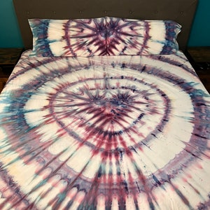 May include: A tie-dye bed set featuring a spiral design in shades of white, burgundy, and navy blue. The set includes a fitted sheet, a flat sheet, and two pillowcases. The bedding is on a bed with a gray headboard.