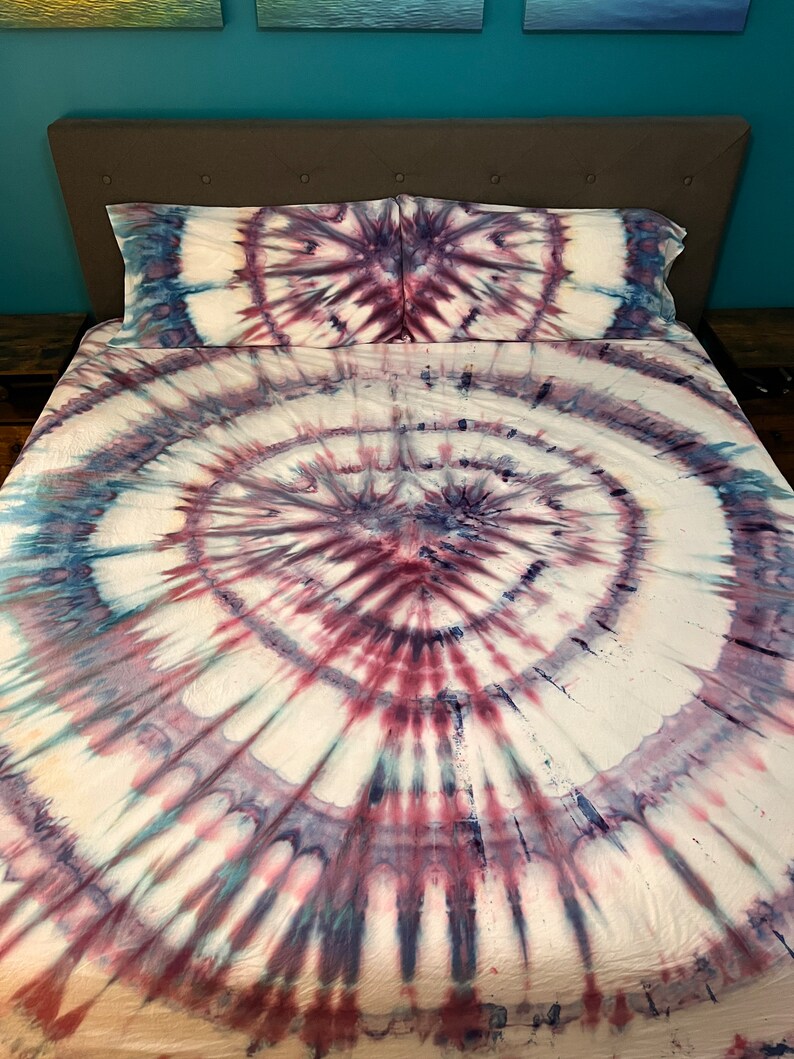 May include: A tie-dye bed set featuring a spiral design in shades of white, burgundy, and navy blue. The set includes a fitted sheet, a flat sheet, and two pillowcases. The bedding is on a bed with a gray headboard.