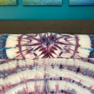 May include: A tie-dye bedding set featuring a white base with purple and blue spiral patterns. The set includes two pillowcases and a fitted sheet, creating a vibrant and artistic look for the bedroom. The fabric appears soft and comfortable.