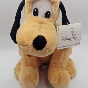 May include: A plush Pluto dog toy with soft yellow fur, long black ears, and a black nose. The toy features a white tag attached to the side that reads Disneyland Paris Collection exclusive Parcs et Disney Stores with a castle illustration.