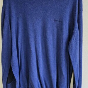 May include: A blue long-sleeve sweater with a crew neckline, featuring the text AJ Armani Jeans on the inner collar label and the Armani Jeans logo embroidered on the chest. The sweater has dark elbow patches and is displayed on a black hanger.