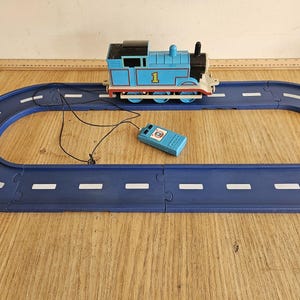 Puede incluir: Un tren de juguete azul de Thomas y sus amigos con el número 1 en el lateral, conectado por un cable negro a un mando a distancia azul. El tren se sitúa sobre una vía ovalada de plástico azul con líneas blancas, de 76 cm de largo.