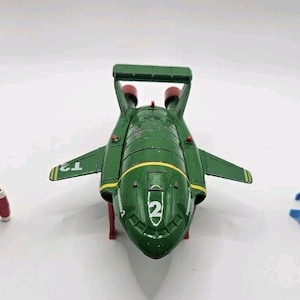 Puede incluir: Tres vehículos de juguete: un cohete rojo con detalles negros, un avión verde con rayas amarillas y el número 2, y un cohete azul y negro. Sobre un fondo blanco.