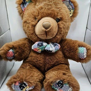 May include: A brown plush teddy bear featuring a patchwork fabric pattern on its ears, bowtie, paw pads, and foot pads. The patchwork fabric consists of various small floral and geometric prints in shades of blue, green, yellow, and red.