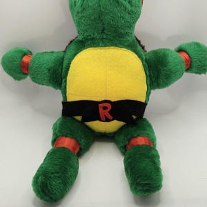 May include: A plush green turtle toy featuring a bright orange eye mask, matching orange arm and leg bands, and a yellow chest plate. A black belt is wrapped around the waist, featuring a prominent red letter R embroidered on the front center.